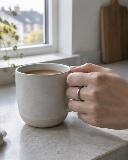 A hand holding a mug of tea by a kitchen window, wearing a titanium smart ring